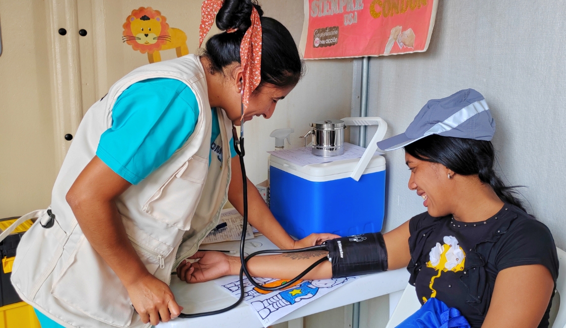 Profesional de Prisma ONG toma la presión arterial a una mujer migrante en un punto de atención del proyecto ECHO ASTRAL en Tumbes, fortaleciendo la salud primaria y el bienestar comunitario.